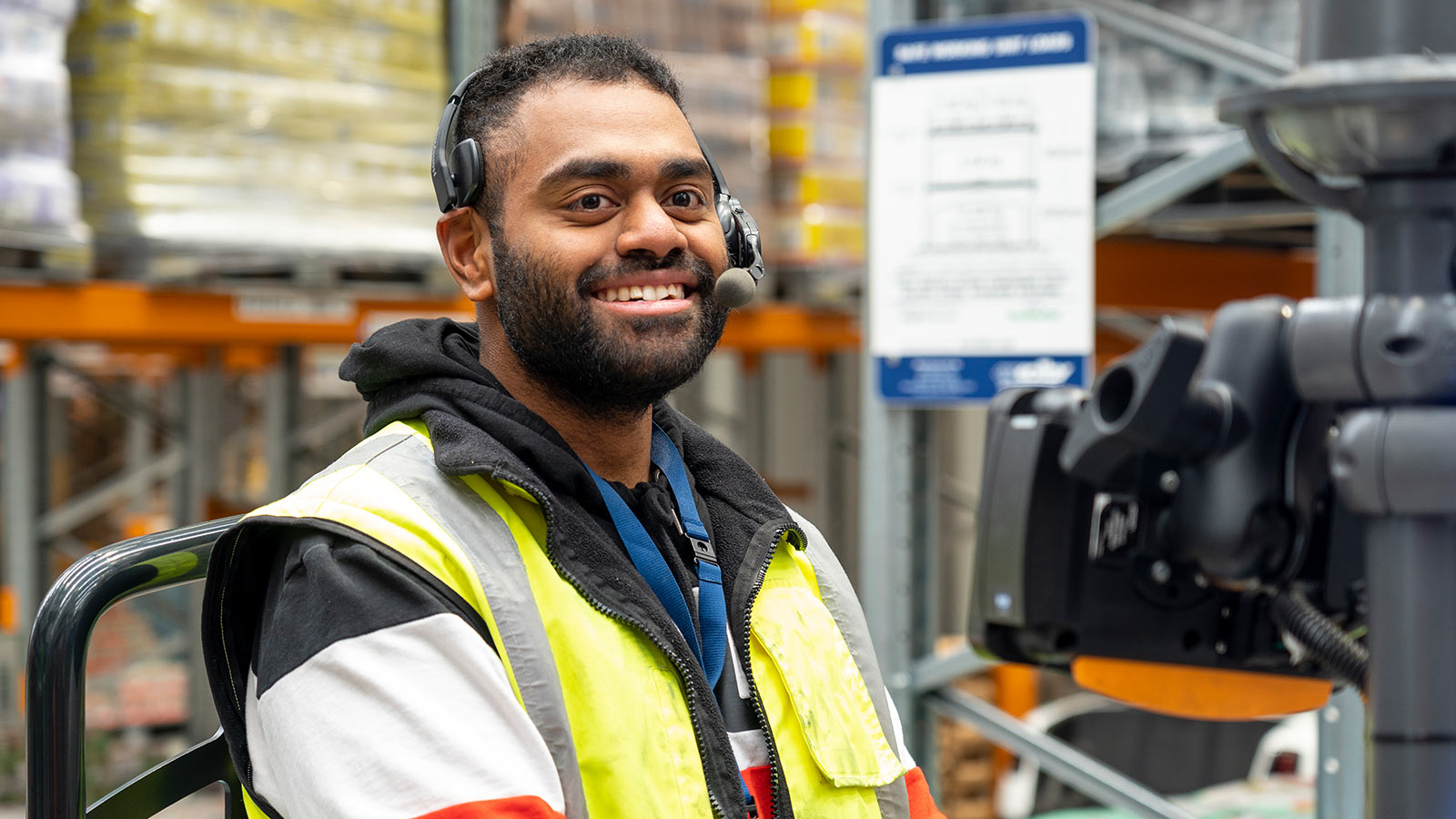 A male warehouse operator smiles as he wears his Voice Picking headset at work, standing on his pallet moving vehicle.