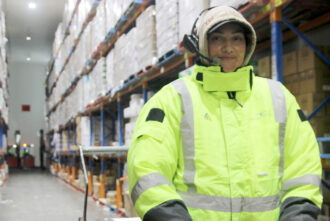 A female warehouse operator wearing a Voice Picking headset and high-visability clothing within an aisle of pallet racking.